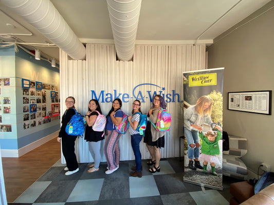 Five women pose holding colorful backpacks in front of a "Make A Wish" wall sign and a Western Chief banner.