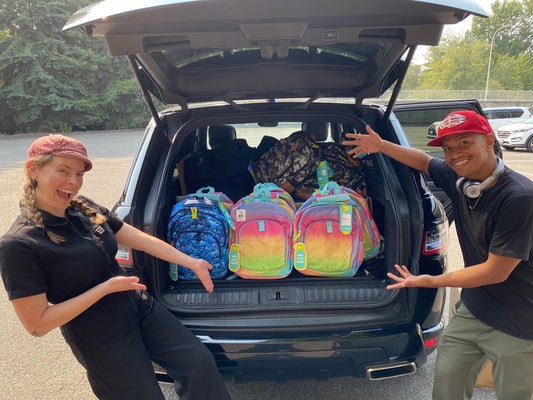 Two people point to backpacks filling the open trunk of an SUV in a parking lot.
