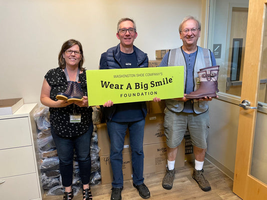 Three people hold a Wear A Big Smile Foundation sign in an office, one holding a pair of boots and another holding a pair of sandals, with shrink-wrapped pallets behind them.