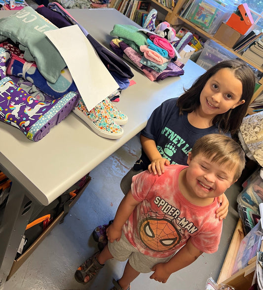 A girl and a young boy in a Spider-Man shirt stand smiling beside a table of folded children's clothing and shoes in a boutique shop.