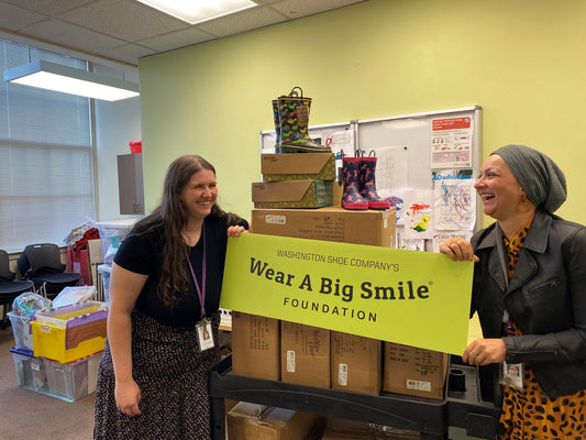 Two women hold a Wear A Big Smile Foundation sign beside a rolling cart stacked with shoe boxes, in an office with a whiteboard and stacked rain boots.