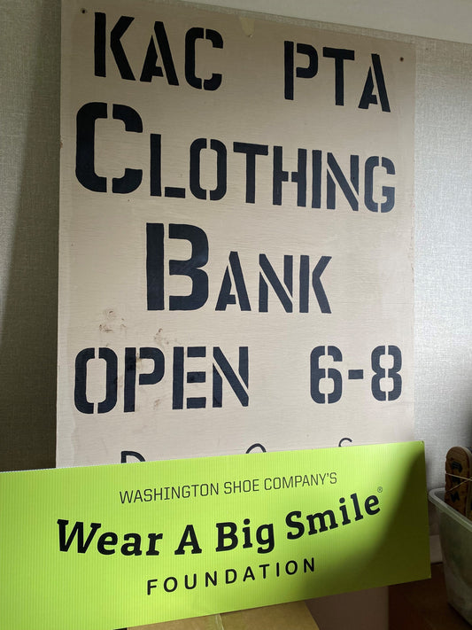 A handmade "KAC PTA Clothing Bank, Open 6-8" sign propped against a wall above a Wear A Big Smile Foundation sign.