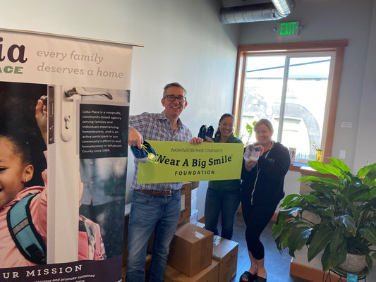 A man and two women hold a Wear A Big Smile Foundation sign beside a Lydia's Place banner and stacked boxes in an office.