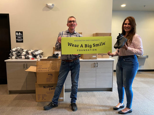 A man and a woman hold a Wear A Big Smile Foundation sign in an office lobby, the woman holding a pair of black boots, with bagged shoes stacked on a counter.