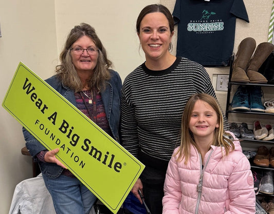 Two women hold a Wear A Big Smile Foundation sign while a young girl in a pink jacket stands nearby, in front of a shoe rack and a Sunrise Elementary t-shirt on the wall.