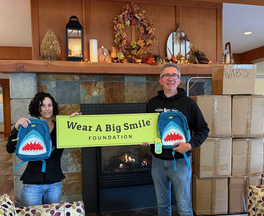 A man and a woman hold shark-themed backpacks and a Wear A Big Smile Foundation sign in front of a fall-decorated fireplace mantel, with stacked donation boxes beside them.