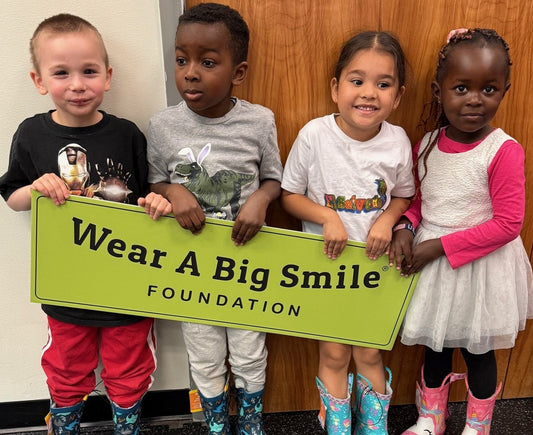 Four young children hold a Wear A Big Smile Foundation sign together, each wearing a different pair of colorful rain boots.