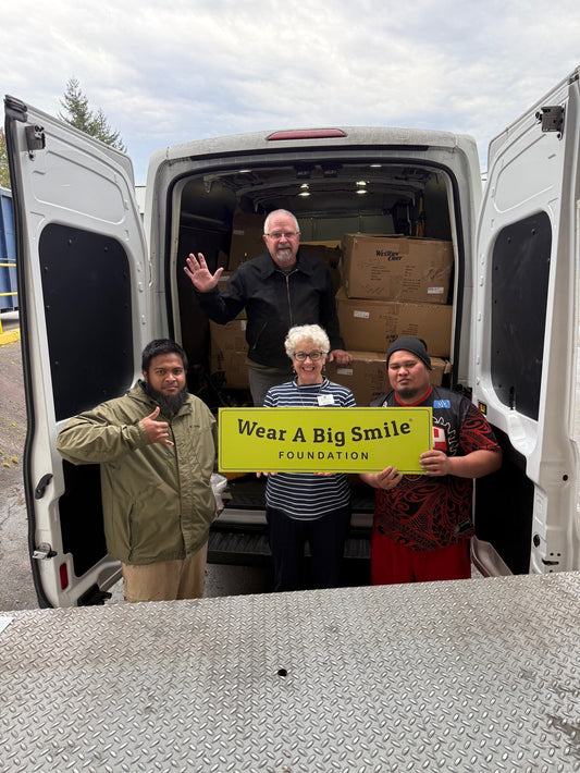 Four people stand in the open cargo area of a van packed with Western Chief shoe boxes, holding a Wear A Big Smile Foundation sign.