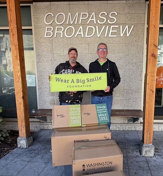 Two men hold a Wear A Big Smile Foundation sign in front of a "Compass Broadview" sign, with stacked Western Chief and Washington Shoe Company boxes at their feet.