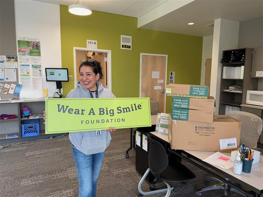 A woman holds a Wear A Big Smile Foundation sign in an office, with stacked Western Chief shoe boxes on a table beside her.