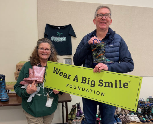 A woman holds a pink glitter rain boot and a man holds a pair of monster-truck print rain boots on either side of a Wear A Big Smile Foundation sign, with rows of children's shoes displayed on a table behind them.