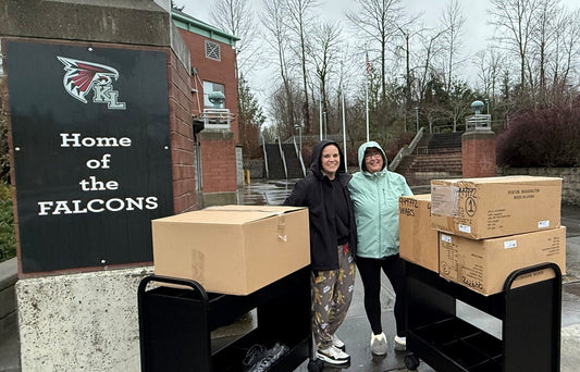 Two women in rain jackets stand beside carts of boxes in front of a "Home of the Falcons KL" sign outside a school.