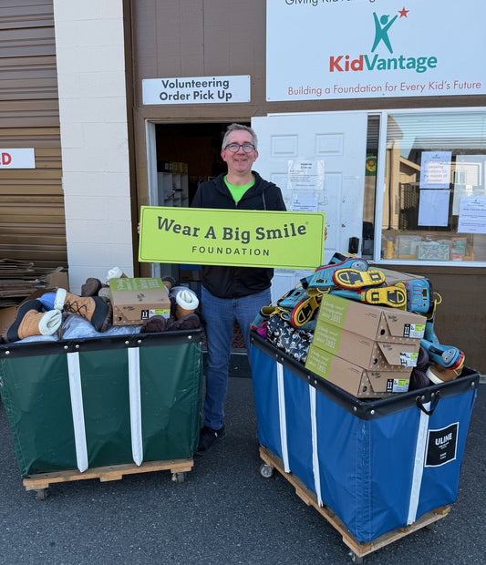 A man holds a Wear A Big Smile Foundation sign in front of a KidVantage building entrance, beside two large bins overflowing with donated shoes and boxes.