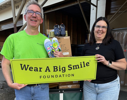 A man and a woman hold a Wear A Big Smile Foundation sign in front of stacked Western Chief shoe boxes, the man holding a pair of rainbow-striped children's shoes.