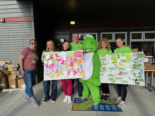 Six people and a green dinosaur mascot pose outside a building entrance, two of them holding poster boards covered with colorful paper boot and dinosaur cutouts signed by kids.
