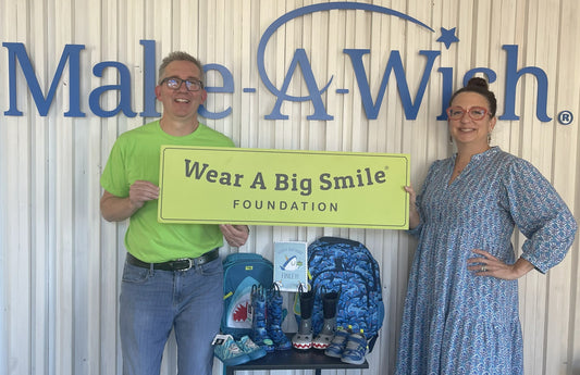 A man and a woman hold a Wear A Big Smile Foundation sign in front of a large Make-A-Wish logo, with shark-themed backpacks, rain boots, and a birthday card for Finley displayed nearby.