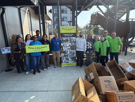 A large group of volunteers poses with a Wear A Big Smile Foundation sign and banner under a covered outdoor area, with stacked shoe boxes in the foreground.