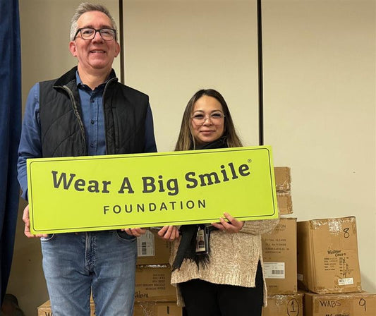 A man and a woman hold a Wear A Big Smile Foundation sign in front of stacked donation boxes marked with numbers.