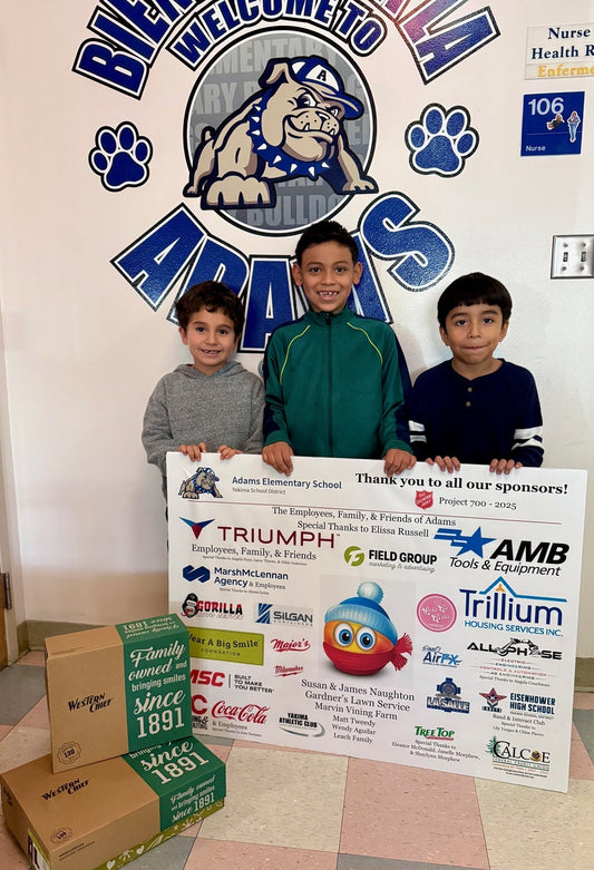 Three young boys stand in front of an Adams Elementary School Bulldogs mural holding a sponsor thank-you banner for Project 700, with Western Chief shoe boxes at their feet.