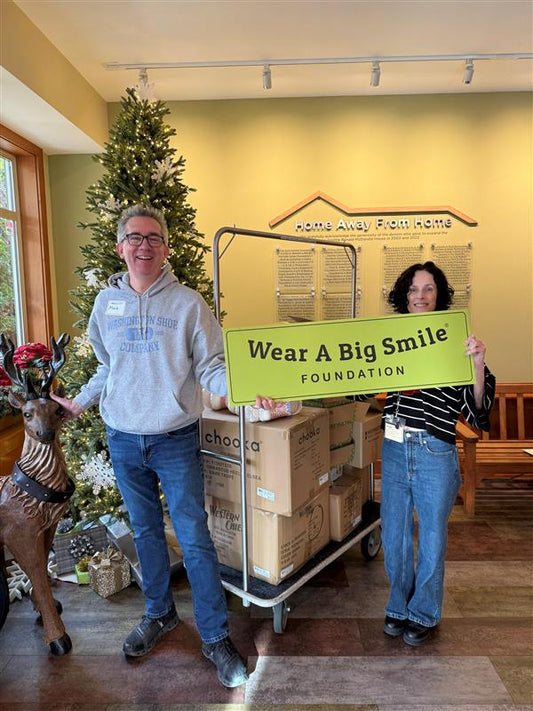 A man and a woman hold a Wear A Big Smile Foundation sign beside a cart stacked with Chooka and Western Chief shoe boxes, in front of a decorated Christmas tree and a "Home Away From Home" plaque.