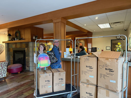 A man and a woman hold backpacks in the Ronald McDonald House lobby beside a cart of Western Chief boxes marked “Done,” with a stone fireplace behind them.