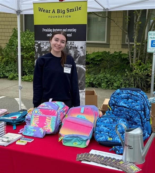 A young woman stands at a Wear A Big Smile Foundation table under a tent, displaying colorful backpacks for an open house event.