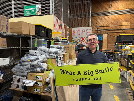 A man holds a Wear A Big Smile Foundation sign in a warehouse storage area stacked with donated shoe boxes and bins, with a colorful "HOPE" sign on the wall behind him.