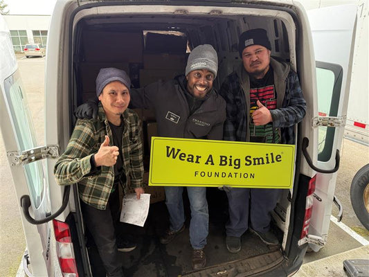 Three men pose in the open back doors of a delivery van holding a Wear A Big Smile Foundation sign, surrounded by stacked boxes.