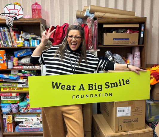 A woman poses playfully making a peace sign behind a Wear A Big Smile Foundation sign, in a classroom storage area filled with board games, books, and backpacks.