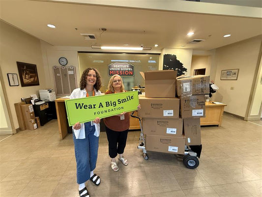 Two women hold a Wear A Big Smile Foundation sign in front of a “Seattle's Union Gospel Mission” sign, with a hand cart stacked with boxes beside them.