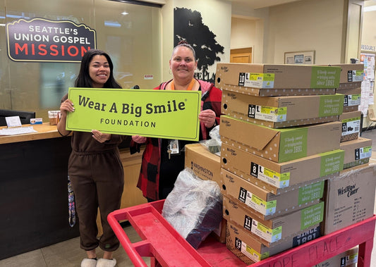 Two women hold a Wear A Big Smile Foundation sign at the Seattle's Union Gospel Mission reception desk, with stacked Western Chief shoe boxes on a cart beside them.
