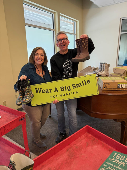 A man and a woman hold a Wear A Big Smile Foundation sign in front of a “Seattle's Union Gospel Mission” sign, each holding a colorful backpack.