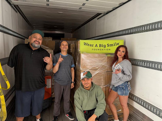 Four men pose in the back of a box truck holding a Wear A Big Smile Foundation sign, surrounded by shrink-wrapped pallets of boxes.