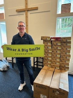 A man holds a Wear A Big Smile Foundation sign next to stacked shoe boxes, with a wooden cross visible in the background.