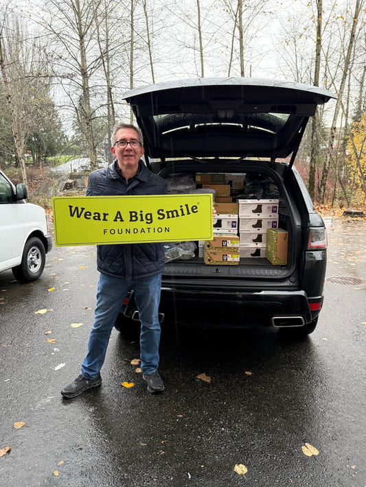 A man holds a Wear A Big Smile Foundation sign in front of an SUV's open trunk packed with shoe boxes on a rainy day, with bare trees in the background.