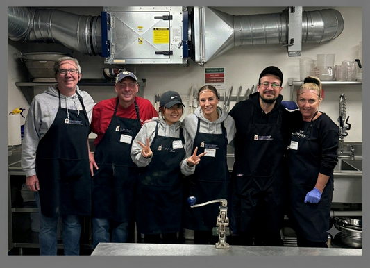 Six volunteers in aprons smiling together in a commercial kitchen after preparing a meal for Ronald McDonald House guests.