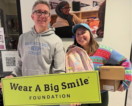 A man and a woman hold a Wear A Big Smile Foundation sign in an office, the woman holding a pink backpack and the man holding a boxed pair of children's rain boots, with a classroom photo on the wall behind them.