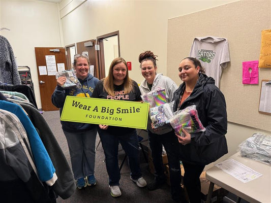 Four smiling women hold a Wear A Big Smile Foundation sign in a clothing bank, two of them holding bagged rain boots, with a Green River Gators t-shirt hanging on the wall behind them.