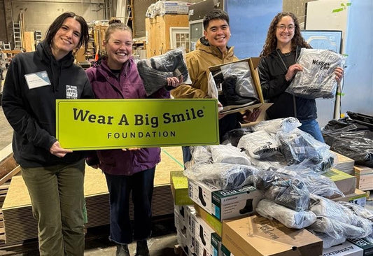 Four people hold a Wear A Big Smile Foundation sign in a warehouse, three of them holding pairs of black boots and shoes, with stacked boxes and shelving in the background.