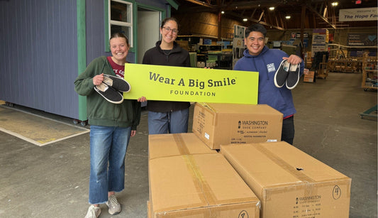 Four smiling people pose together in front of a community resource bulletin board, holding pairs of pink camo, black, and sparkly pink rain boots alongside a Wear A Big Smile Foundation sign.