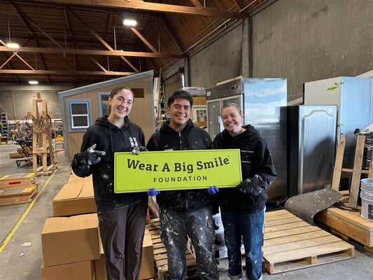 Three paint-splattered volunteers hold a Wear A Big Smile Foundation sign inside a warehouse where tiny houses are being built.