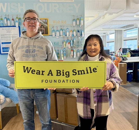 A man and a woman hold a Wear A Big Smile Foundation sign in front of a "Wish Journey" wall covered with colorful wish tags.