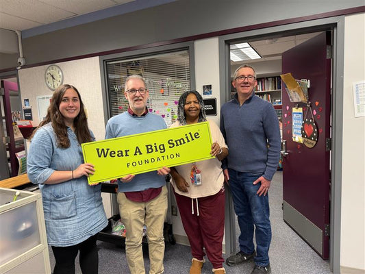 Four smiling people stand together holding a Wear A Big Smile Foundation sign in a school office decorated with Valentine's Day hearts.