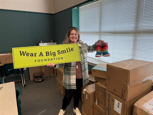 A smiling woman in a plaid shirt holds a Wear A Big Smile Foundation sign and a pair of rainbow-striped rain boots in a classroom, with stacked shoe boxes beside her.