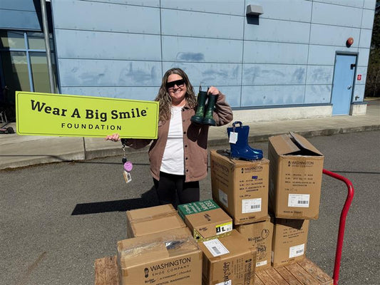 A woman holds a pair of black rain boots and a Wear A Big Smile Foundation sign outside a school building, with more boots and donation boxes stacked on a cart beside her.