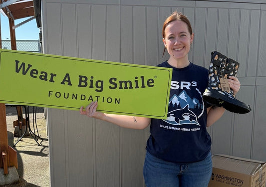 A woman in an SR3 Sealife Response, Rehabilitation, and Research t-shirt smiles while holding a pair of animal-print rain boots, standing beside a Wear A Big Smile Foundation sign.
