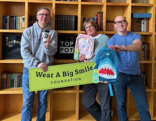 Three people stand among bookshelves holding a Wear A Big Smile Foundation sign, one man holding a Top Pot coffee cup, a woman holding a pink unicorn backpack, and a man holding a blue shark backpack.