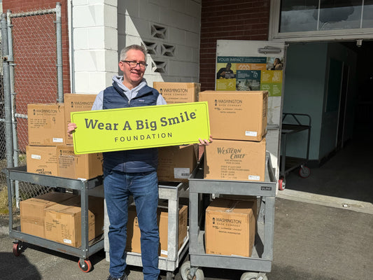 A man in a Patagonia vest holds a Wear A Big Smile Foundation sign in front of stacked Washington Shoe Company and Western Chief boxes outside a brick building.