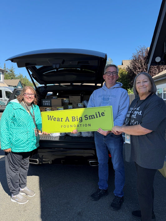 Mark and two people holding a Wear A Big Smile Foundation sign in front of an SUV trunk loaded with donated shoe boxes