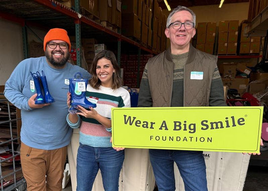 Three people hold a Wear A Big Smile Foundation sign in a warehouse, two of them holding up small blue rain boots.